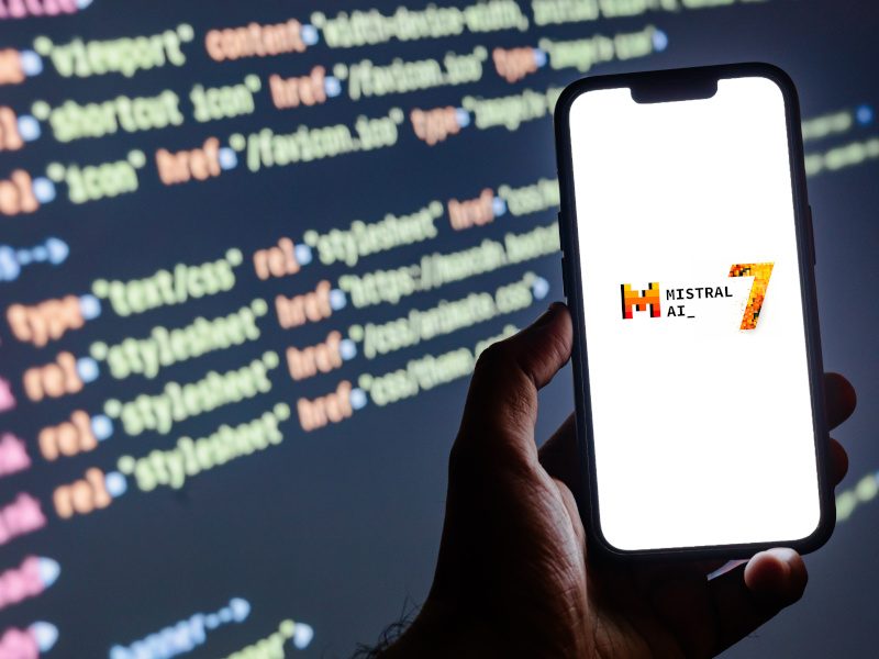 A smartphone with the words ‘MistralAI_7’ present on its screen. In the background, lines of code can be seen on a larger screen.