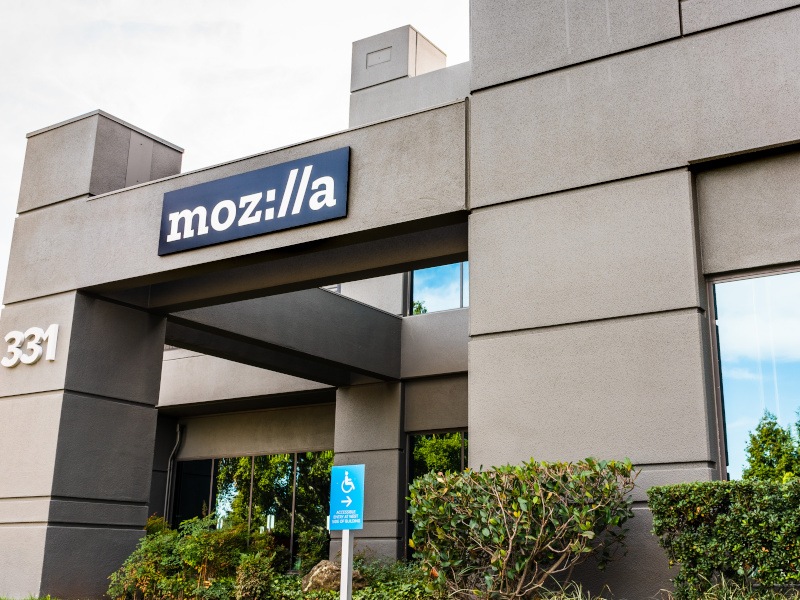 A grey office building with the Mozilla logo on a black sign on top of an archway. The building has grass and shrubbery at the front.