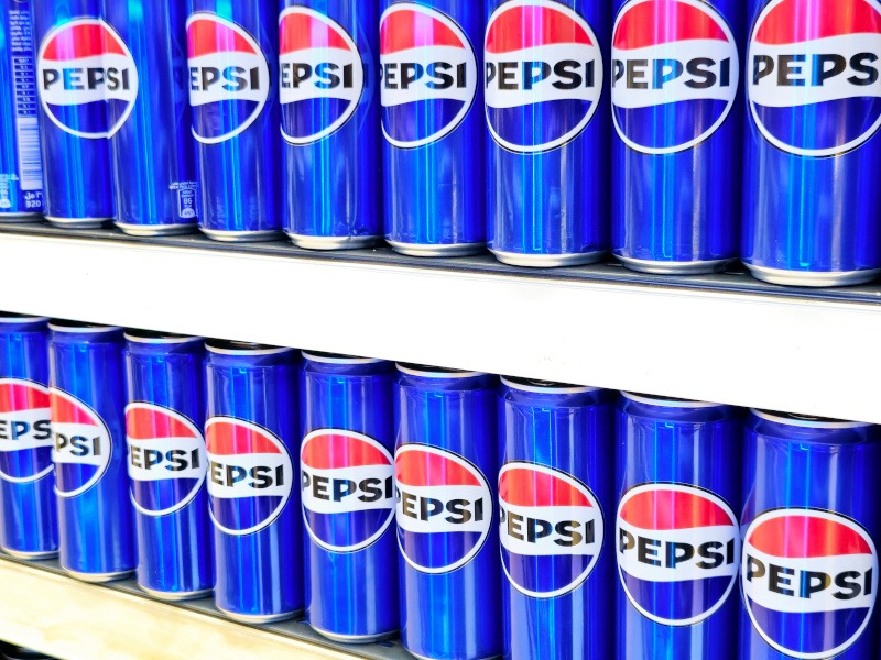 A number of blue metallic cans bearing the blue, white and red Pepsi logo are stacked together on two vertical supermarket shelves.