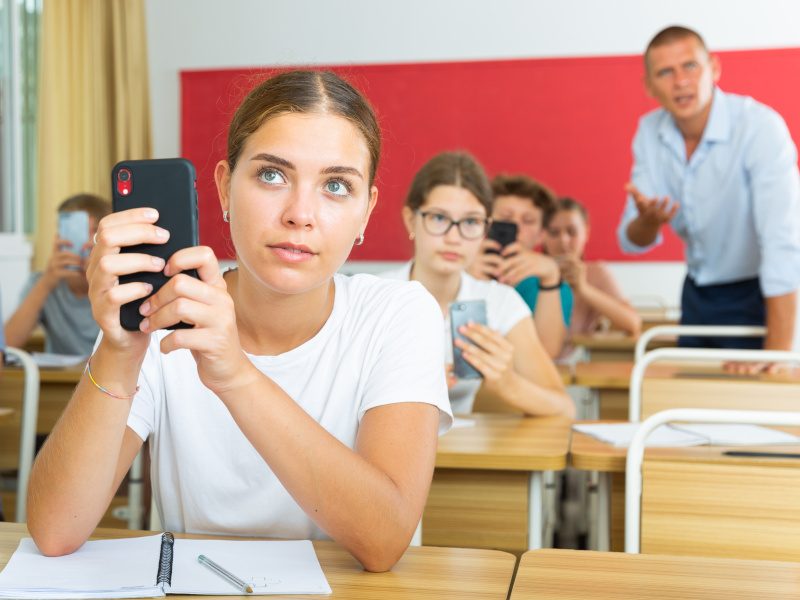 A group of young students in a classroom take out their phones during a lesson. An older man, presumably a teacher, can be seen in the background.