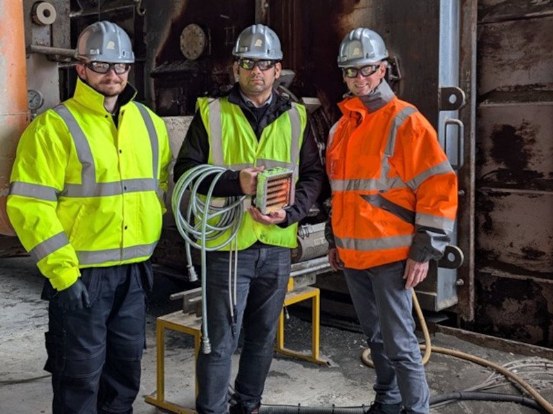 Three men dressed in safety helmets and high visibility jackets gather for a photo which was taken inside a construction facility.