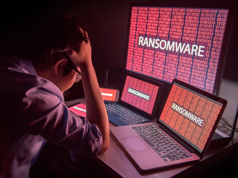 A man appears stressed while he sits at a desk. He is looking at multiple electronic devices displaying the phrase ‘Ransomware’ on their screens.