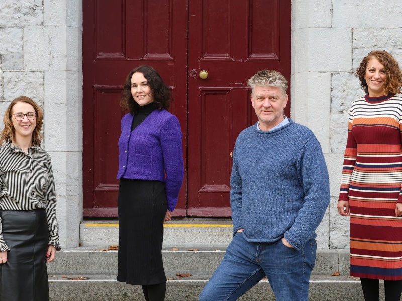 Four people - three women and one man - pose for a photograph in front of a grey-brick building with a red door.