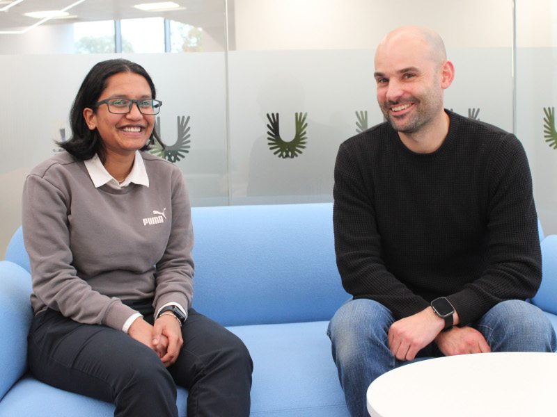 A man and woman sit on a blue couch and smile for a photograph. A frosted glass window can be seen in the background.