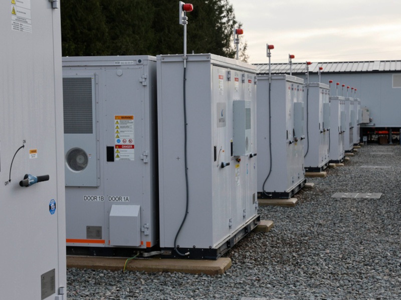 A battery storage site made up of multiple units. There are stones on the ground, and a long building and trees are visible in the background.