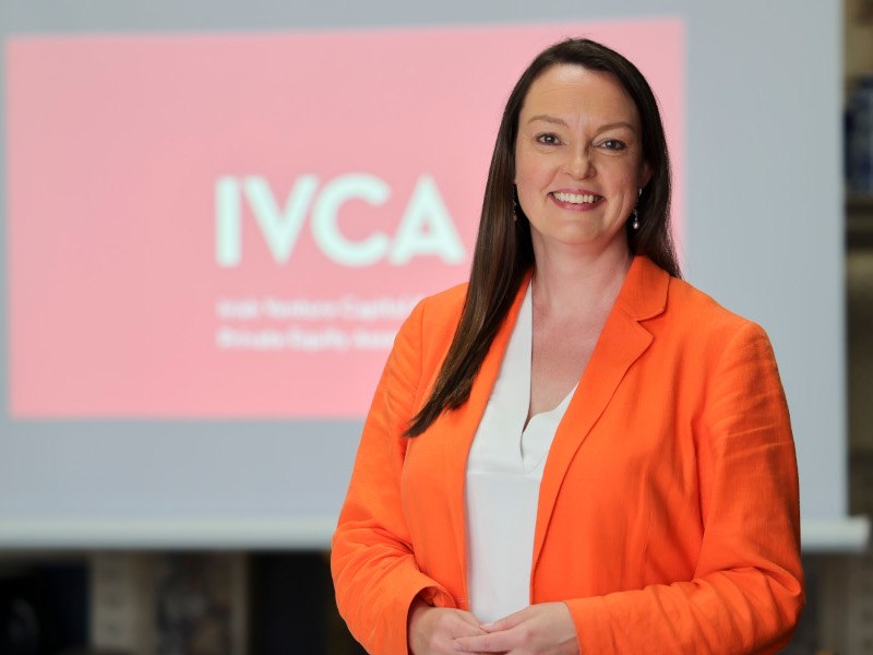 A woman dressed in an orange overshirt smiles while facing the camera. Behind her is a projected image which displays the letters ‘IVCA’.