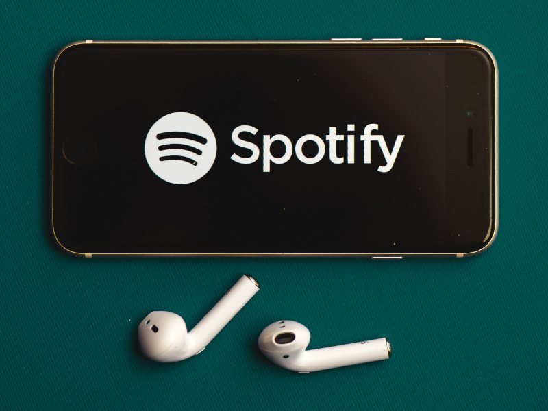 A smartphone against a green background, with the official Spotify logo present on its screen. Two wireless earphones are underneath the phone.