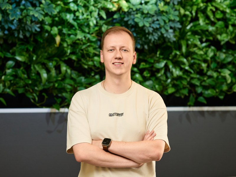 A young man stands in front of a bush while crossing his arms and smiling. He is wearing a light-coloured shirt with the word ‘Storyby’ on it.