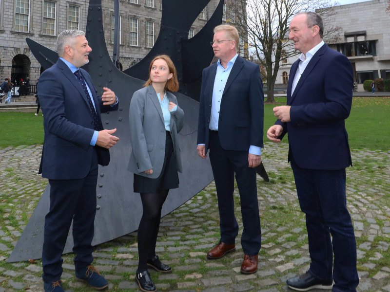 Four people dressed in business attire stand in front of an artistic structure. Grey buildings can be seen in the background.