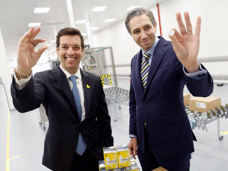 Two men dressed in suits smile for a photograph together in a manufacturing plant. They are both holding up a small capsule in their hands.