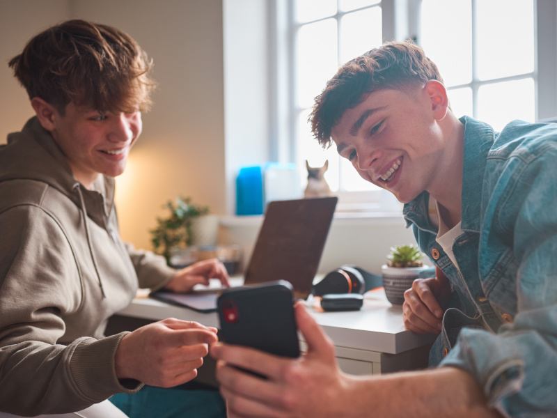 Two teenage boys sit in front of a desk. One of the boys is showing the other something on his phone. They are both smiling.