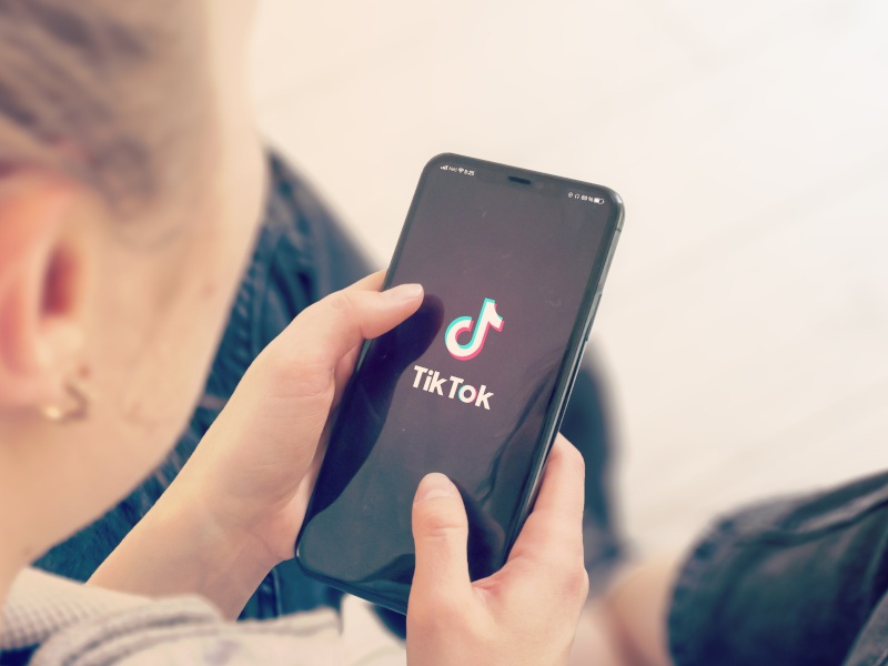 A young person holds a smartphone with the TikTok logo on the screen. The person appears to be wearing a white top and black jeans.