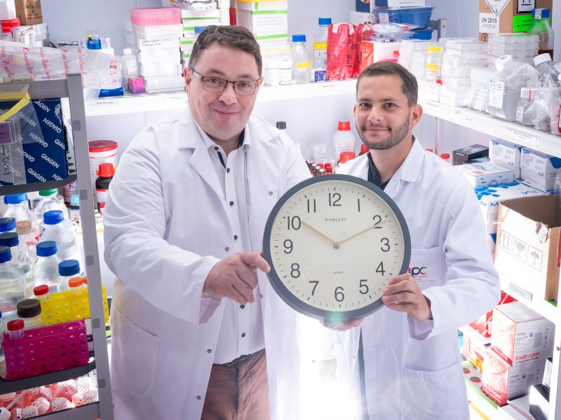 Two people in white lab coats holding a clock.