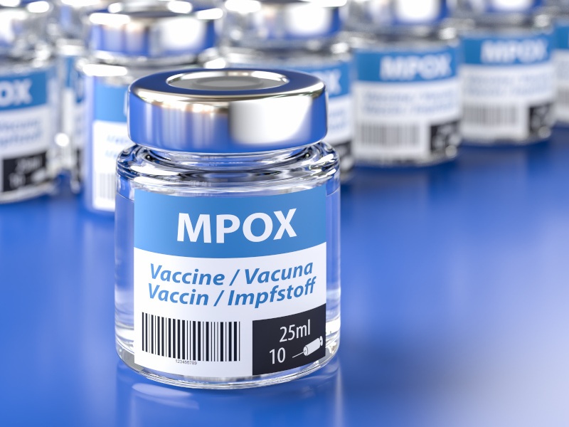 A small glass container with a label displaying that it contains the vaccine for mpox. A number of similar containers can be seen in the background.