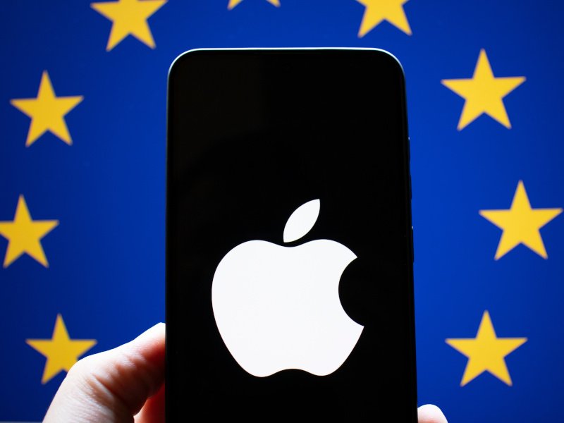 The EU flag in the back with a person holding a smartphone with the Apple logo on it.