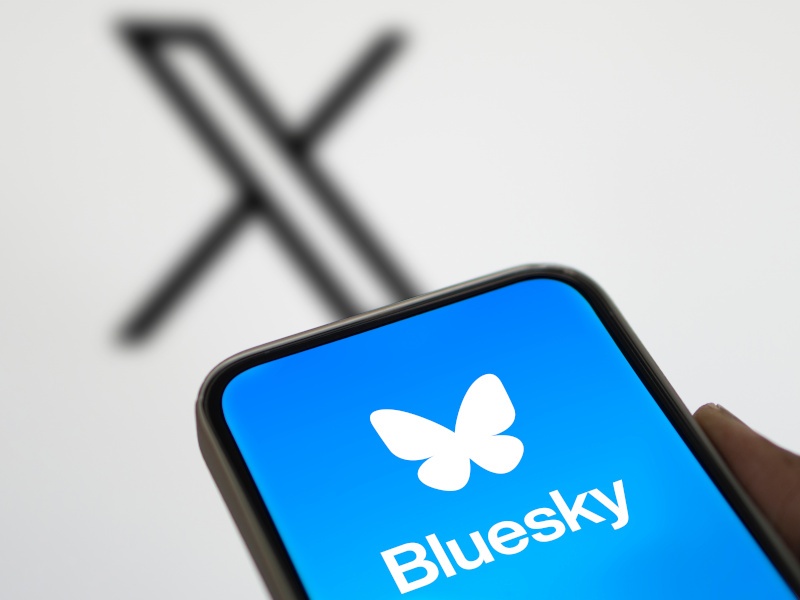 A phone with a large Bluesky logo and the large X logo on a white wall behind.