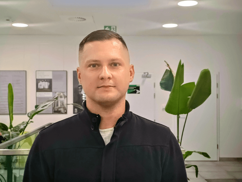 A headshot of Jacek Dąbrowski who wears a black shirt and stands in front of some plants in a wide open office space.