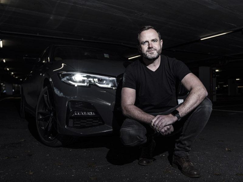 Provizio CEO Barry Lunn crouches in front of a car in a black outfit in a dark space.