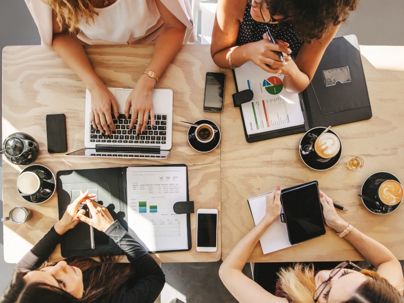 Overhead shot of four people working together on a desk in a brightly lit room.