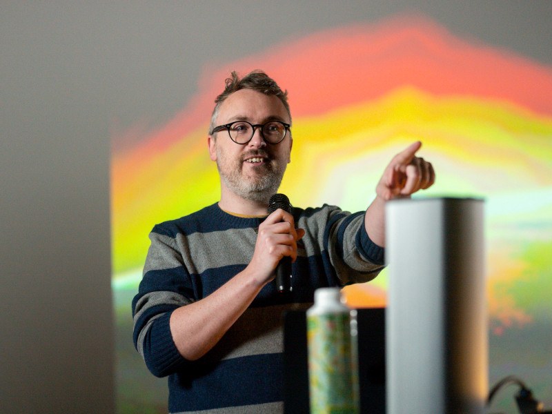 A projector on the back and a man in front of it talking while pointing at something away.