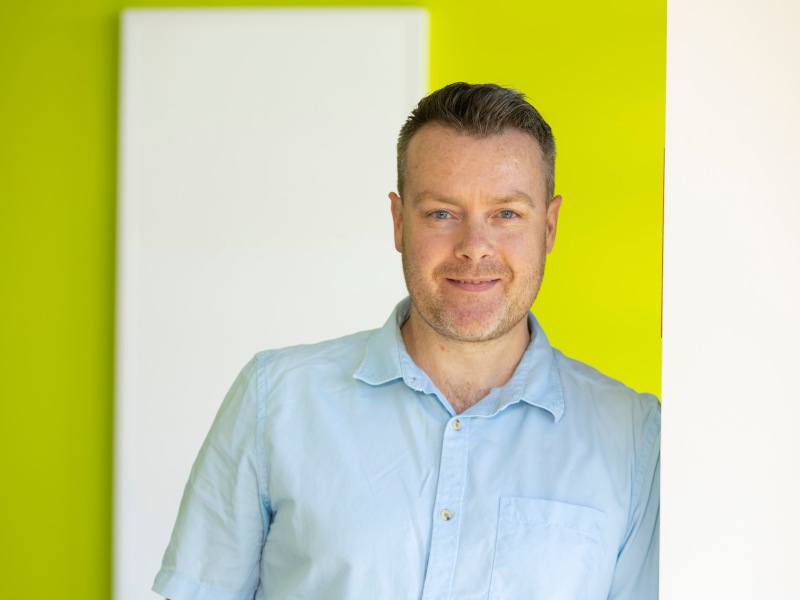 Daniel Hickey leans against a wall wearing a light blue shirt in front of a lime green and white striped wall.