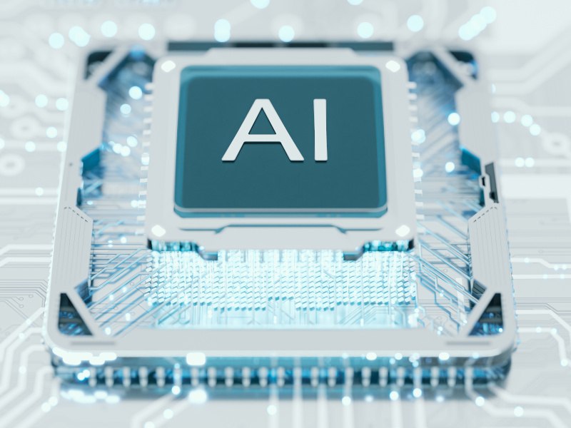 A transparent computer chip with the word ‘AI’ on it. The chip is placed against a transparent circuit board.