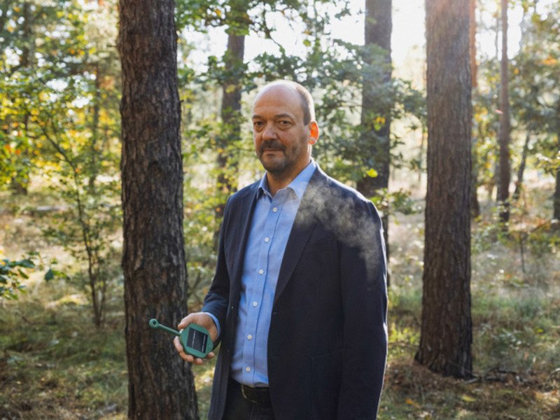 A man wearing a dark suit with a blue shirt stands in a forest setting. He is holding a sensor device in his hand.