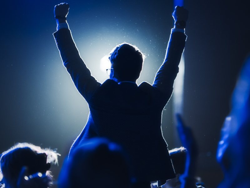 A man wearing glasses and dressed in a suit at an awards ceremony stands up with his arms held out in celebration.