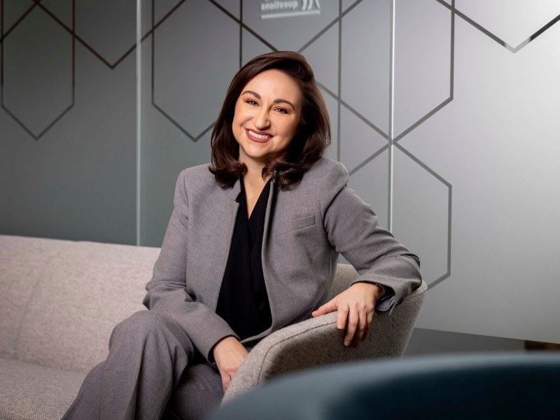 A woman wearing a grey suit, sitting on a couch, smiling.