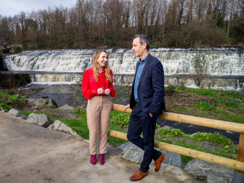 Two people standing against the railing near a river, talking.