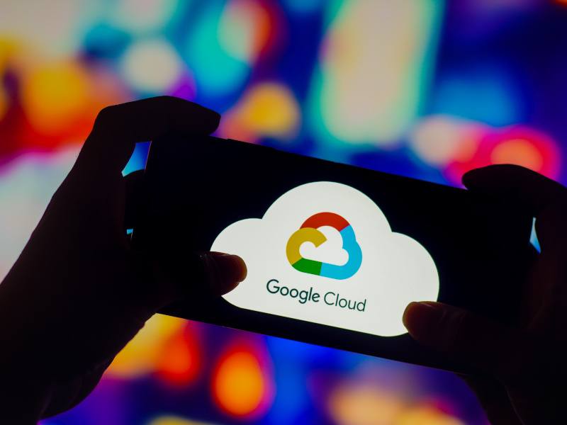 Two hands holding up a smartphone against a blurred background. The Google Cloud logo is displayed on the screen.