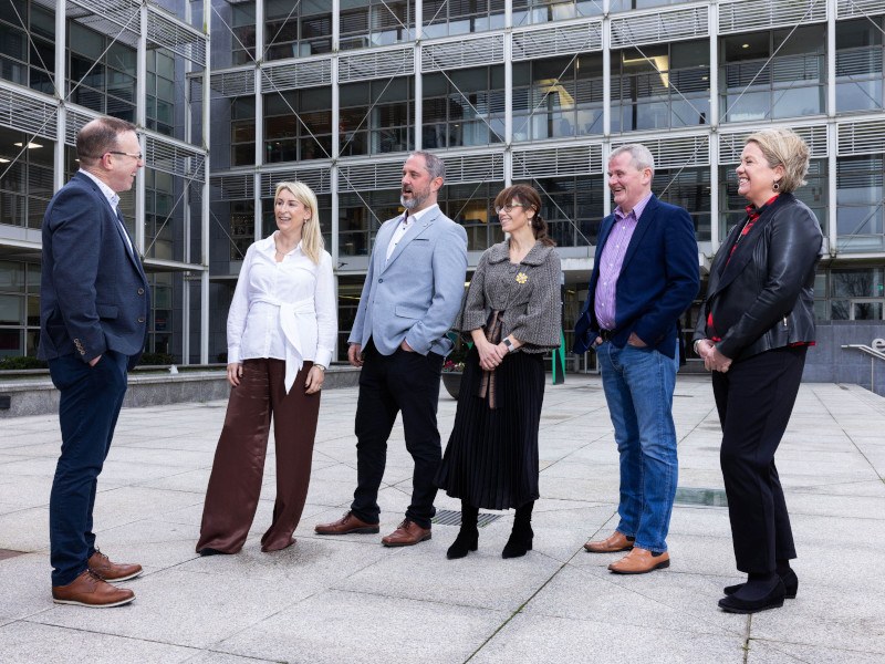 Six people standing outside talking to each other. One man is facing the five others.