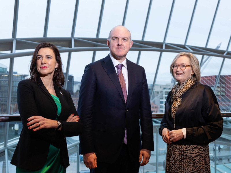 Three business in work attire stand looking to the left of the image with a glass wall behind them.