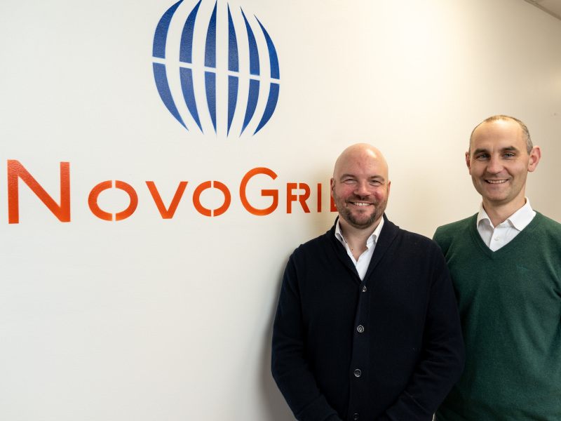 Two men smile for a photograph. On the left-hand side of the photograph, a blank wall with the ‘NovoGrid’ logo can be seen.