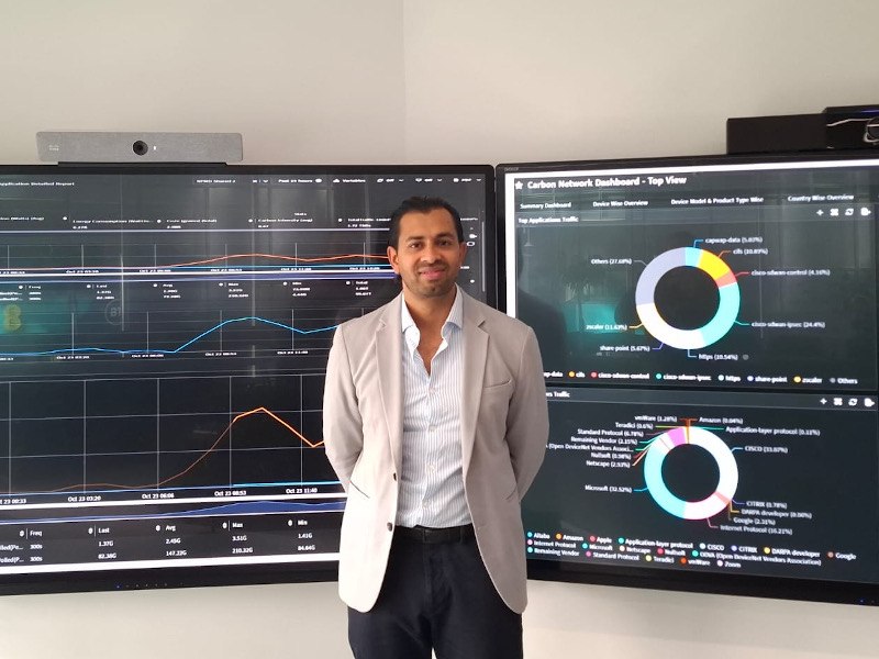 A man wearing a light-coloured blazer stands in front of two large screens with data on them.