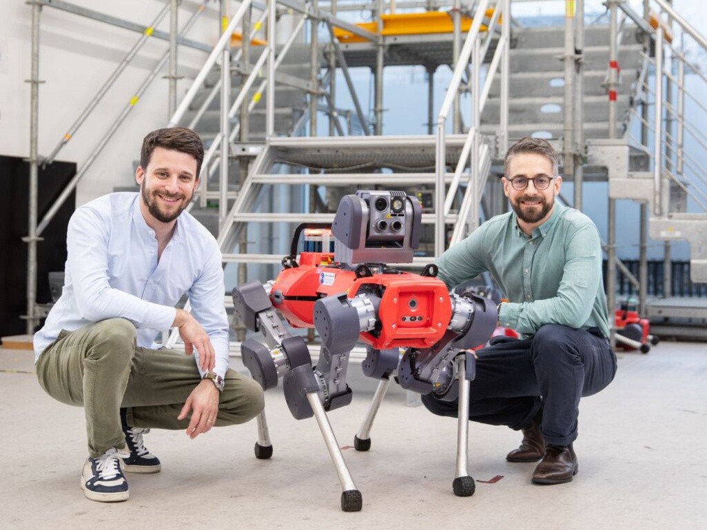 Two men lean down beside a four-legged red robot dog in an industrial setting.