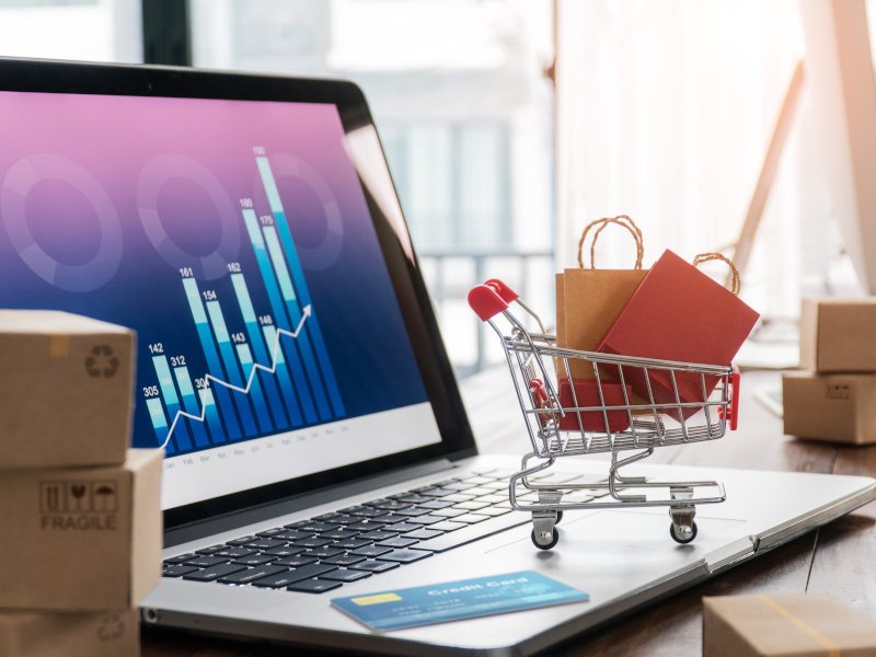 A laptop with a small trolley full of goods, a credit card and cardboard boxes. This image is meant to represent e-commerce.