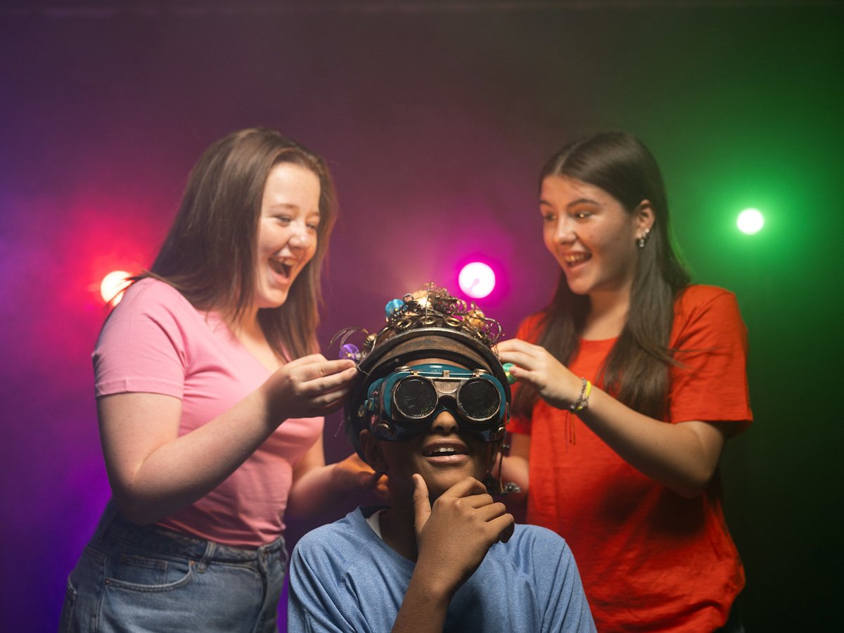 Two young girls stand behind a young boy wearing a futuristic helmet and goggles. There are colourful lights behind them.