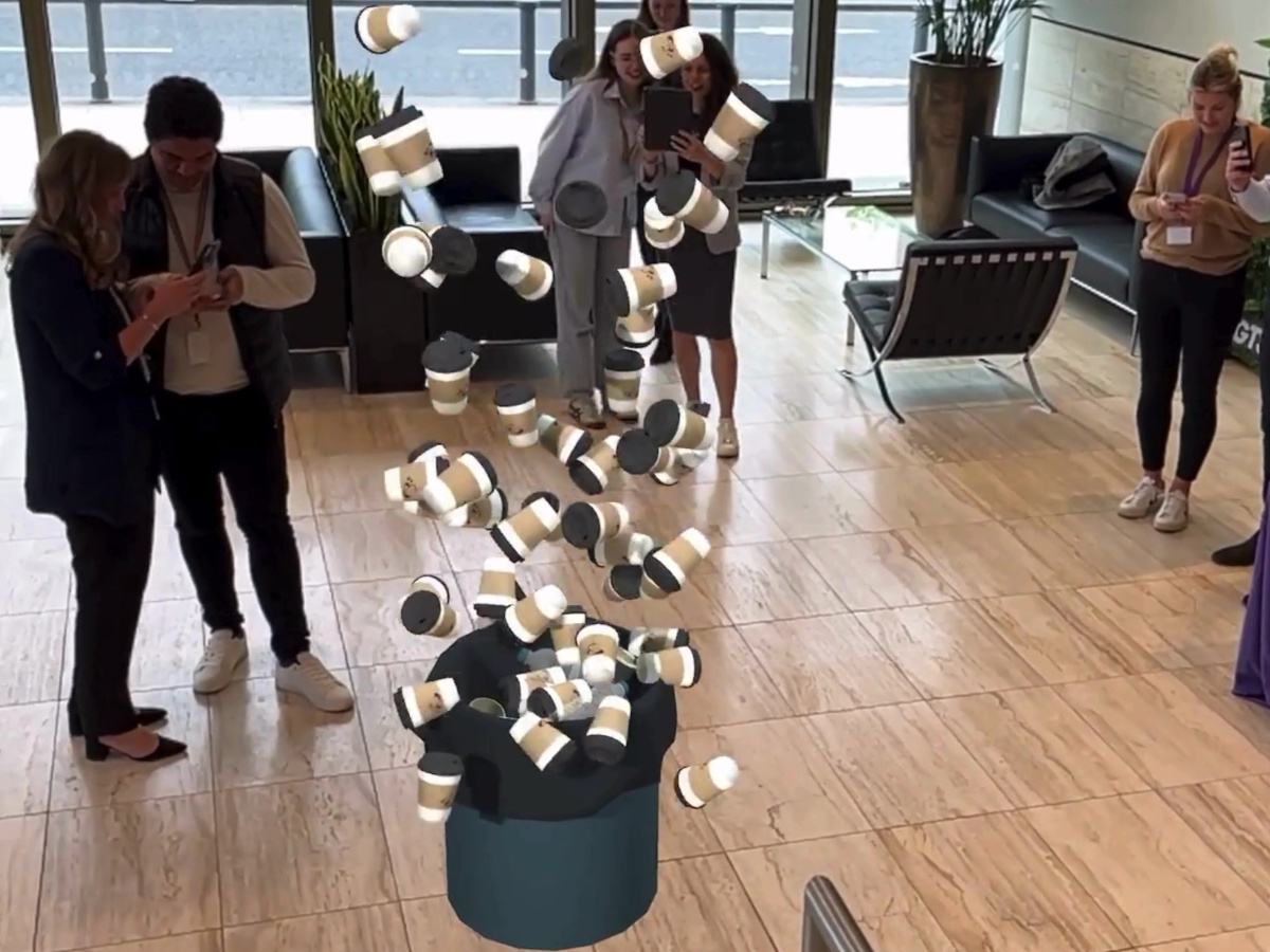 A room of people looking at their phones. In the centre, an AR digital image of a bin overflowing with coffee cups.