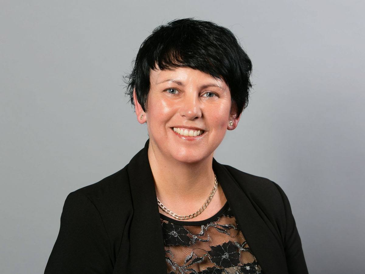 A headshot of a woman with short black hair wearing a black blazer and smiling at the camera against a grey background.