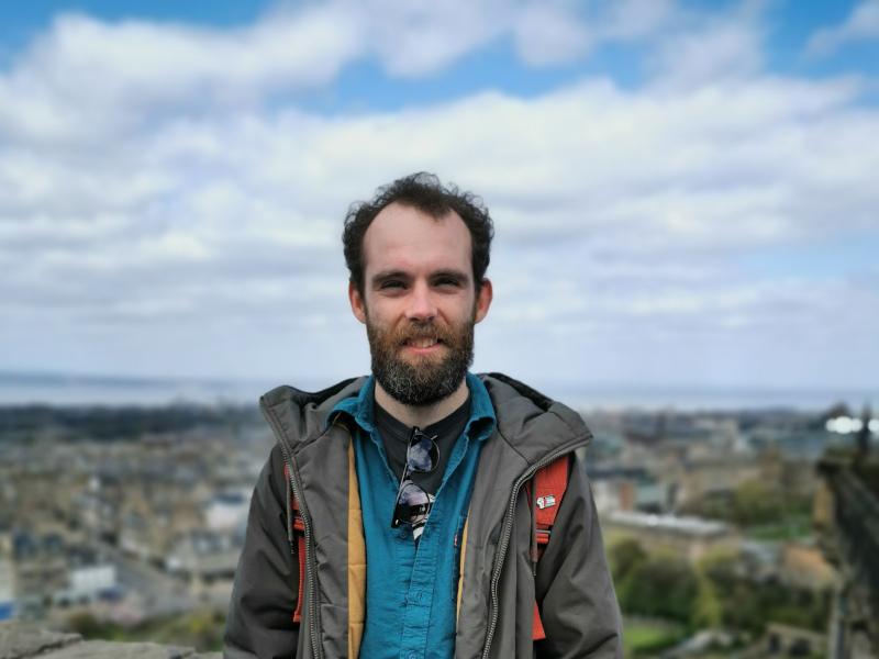 Kevin McDonnell stands on a hill in front of a blurry townscape. He wears a rain jacket and a red backpack.