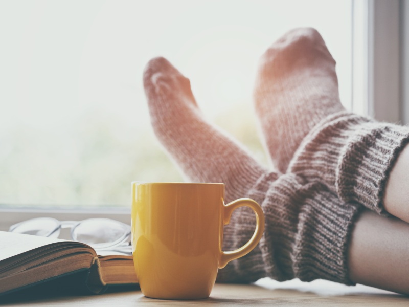 A close-up of a person's feet in comfy-looking woolly socks on a windowsill next to a yellow mug, an open book and a pair of glasses.